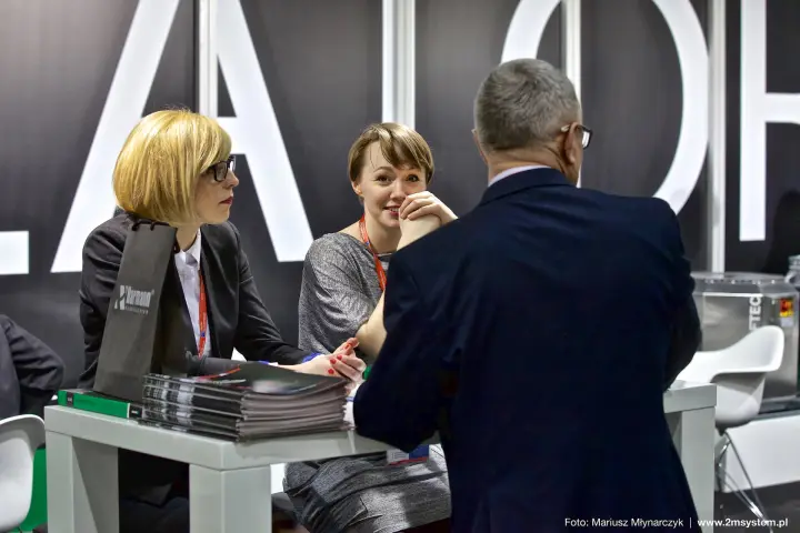 Harmann staff talk with a visitor in front of white lettering at Forum Wentylacja 2016
