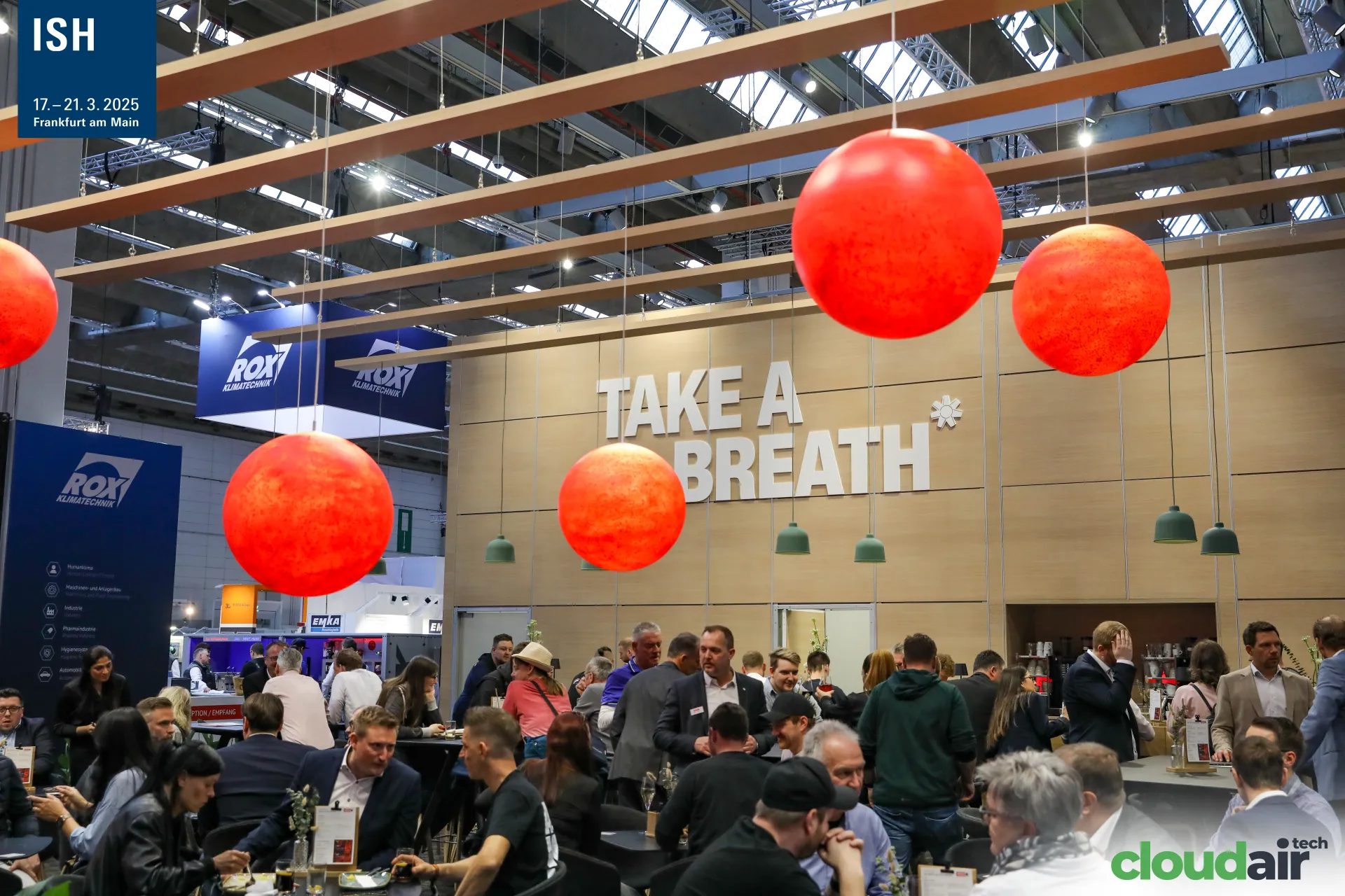 Crowded "TAKE A BREATH" lounge with large orange hanging spheres at ISH 2025