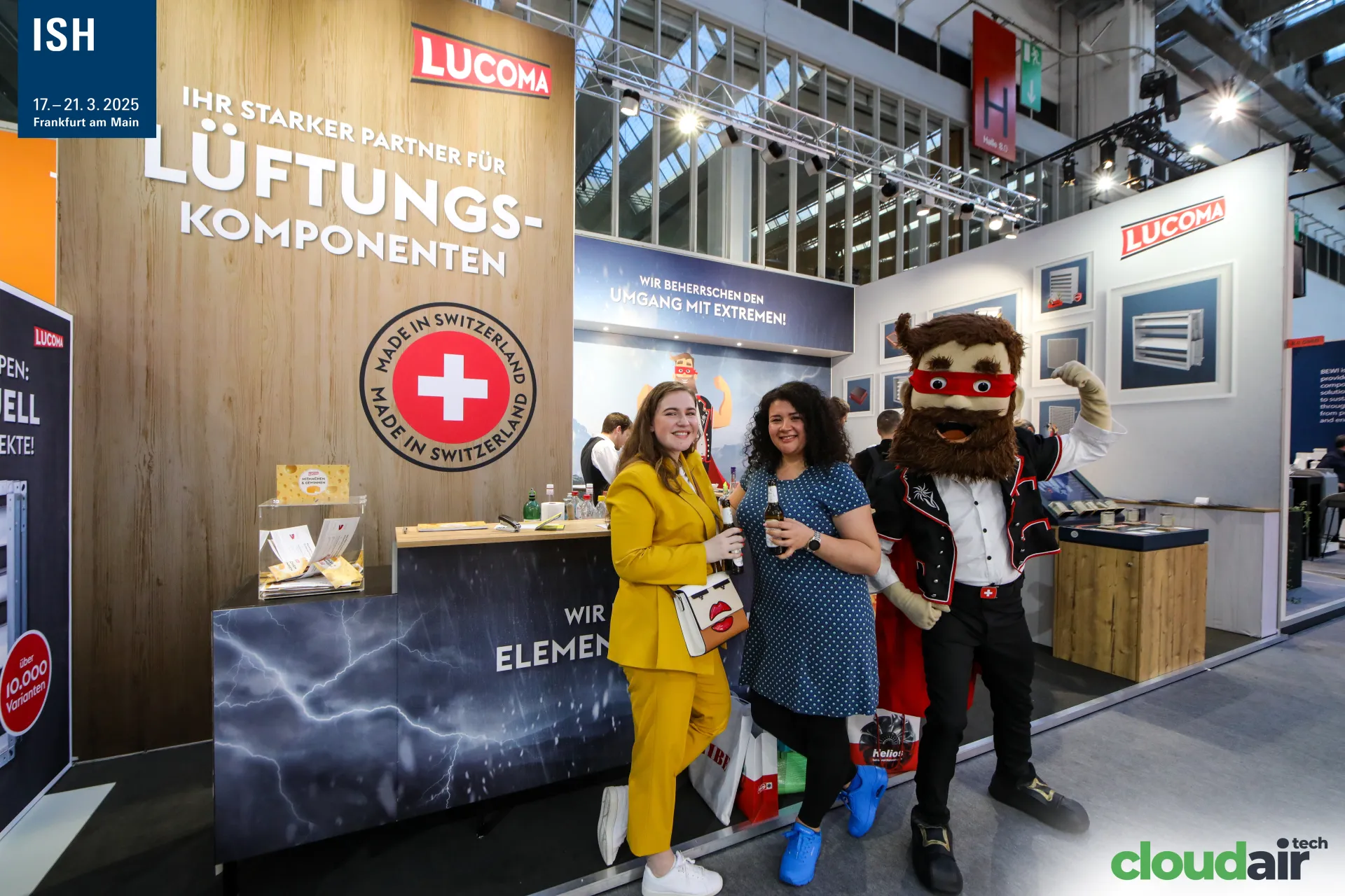 Lucoma mascot flexes beside visitors at the Swiss-made stand at ISH 2025