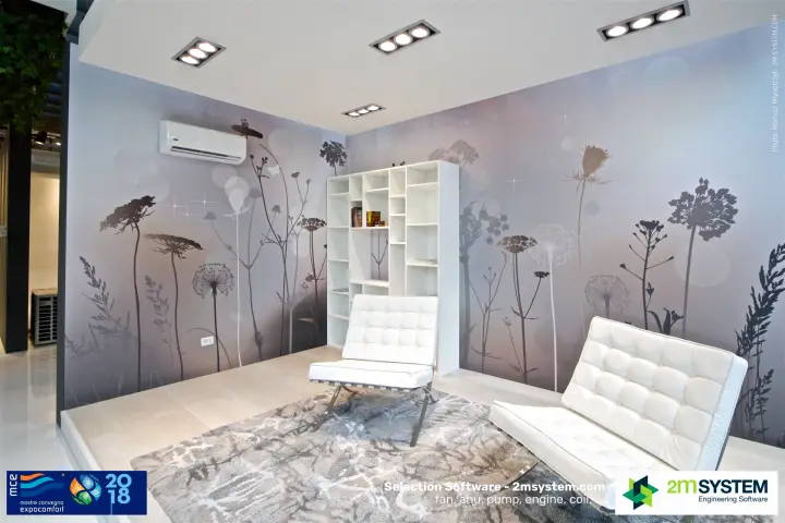 Living-room setting with a wall-mounted split above white tufted lounge chairs at MCE 2018