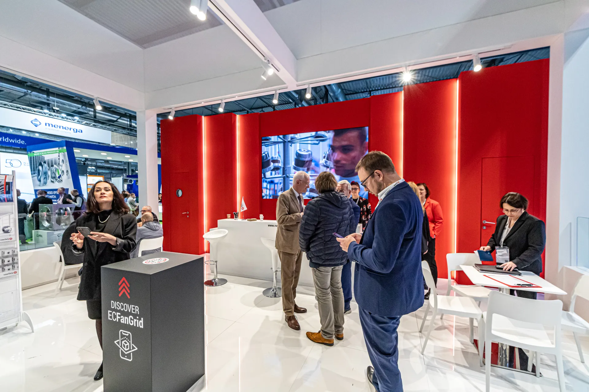 Red-walled stand interior with video screen, ECFanGrid totem and meeting tables at MCE 2024